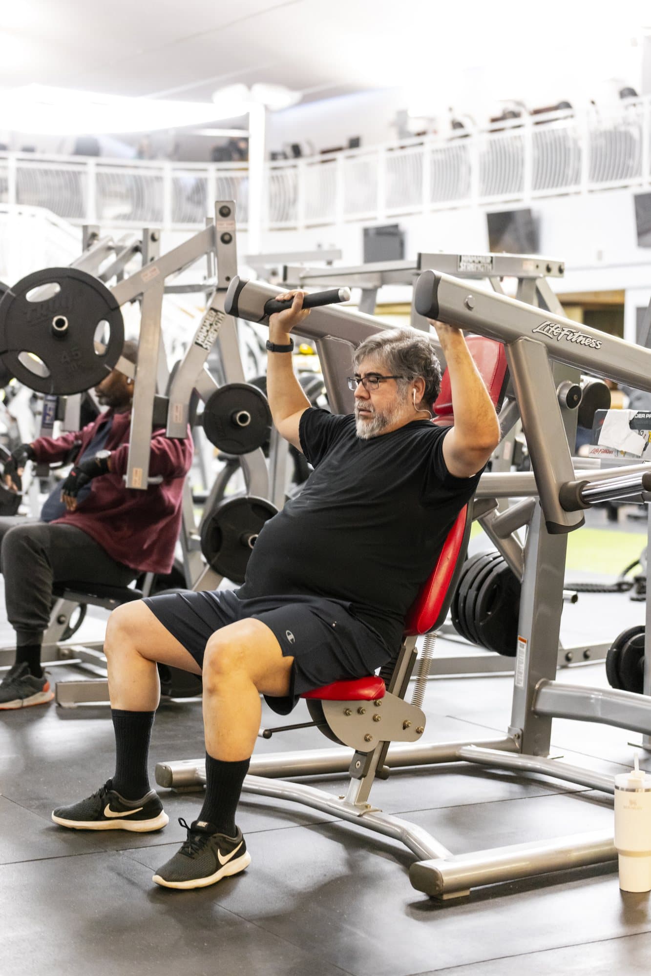 A member presses overhead on a plate-loaded Life Fitness shoulder press while another member trains on a nearby machine on a busy gym floor at an existing W.O.L.F. location (source photo Mullica Hill-52.jpg).